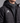 A close-up of a person wearing a black quilted puffer jacket over a dark gray zip-up athletic top. The jacket has a white geometric logo on the left chest and a black hood with drawstrings.