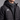 A close-up of a person wearing a black quilted puffer jacket over a dark gray zip-up athletic top. The jacket has a white geometric logo on the left chest and a black hood with drawstrings.