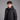 A man wearing a black hooded puffer jacket over a dark gray zip-up sweater, photographed against a solid gray studio background.