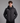 A man wearing a black hooded puffer jacket with a white logo on the left chest, standing against a solid gray studio background.