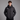 A man wearing a black hooded puffer jacket with a white logo on the left chest, standing against a solid gray studio background.